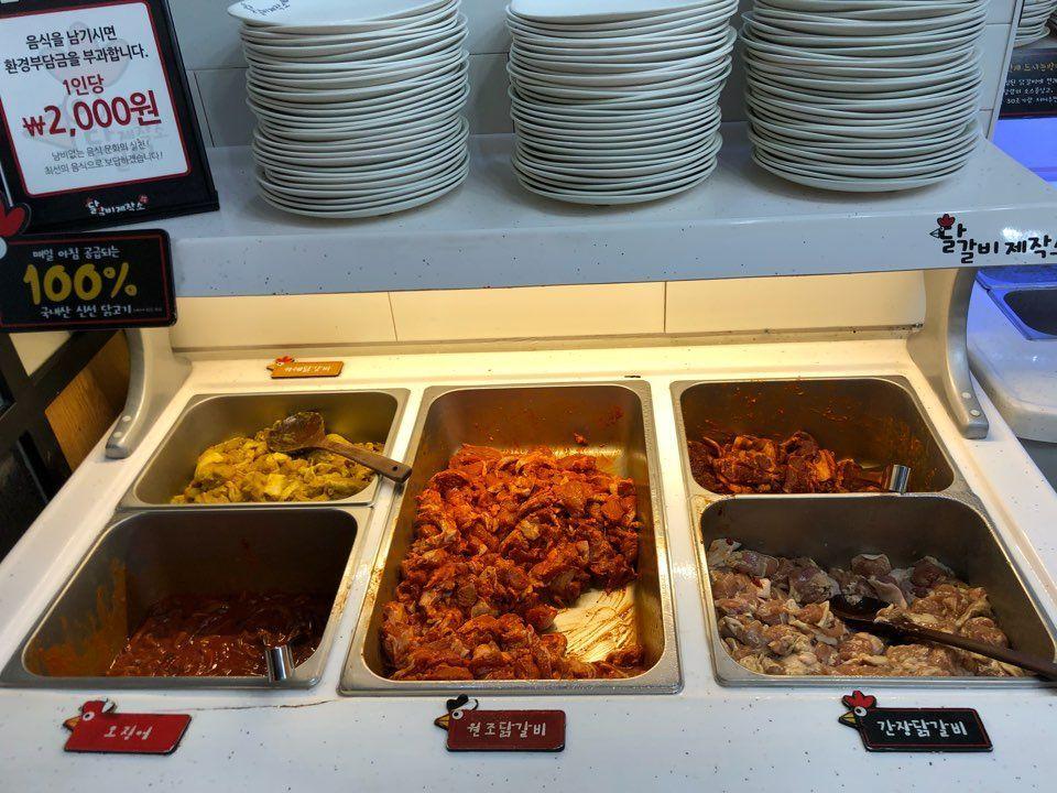 A buffet station offering various flavors of Dakgalbi, with bowls of marinated meats and vegetables under bright lighting, inviting a feast.